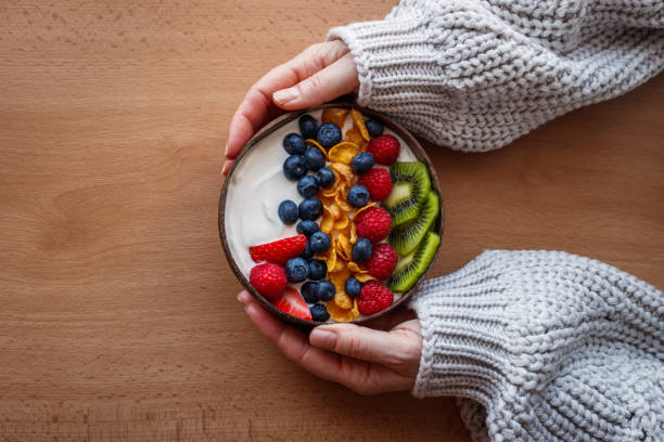 Healthy breakfast in coconut bowl on wooden table Yogurt with corn flakes and blueberry, strawberry, kiwi and raspberry. Womans hand wearing sweater. Vegetarian food probiotic stock pictures, royalty-free photos & images