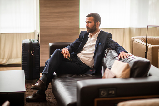 Young businessman seen siting in hotel lobby after finishing a business trip and waiting for a ride on the airport with his luggage packed and ready.