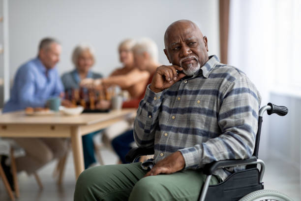 Disabled african american senior man in wheelchair feeling lonely Disabled african american senior man in wheelchair feeling lonely, sitting aside of cheerful elderly men and women playing chess and chatting. Adaptation at retirement house concept common room stock pictures, royalty-free photos & images