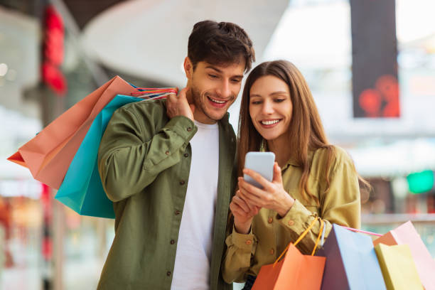 compras en pareja usando la aplicación del teléfono sosteniendo bolsas de comprador en el centro comercial - mercancía fotografías e imágenes de stock