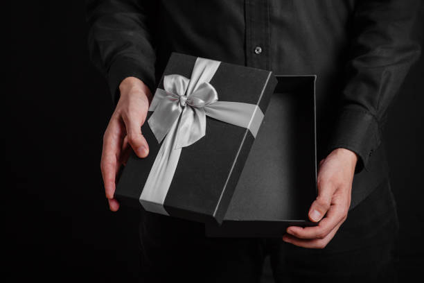istock A guy in a black shirt on a black background holds an open gift box with a silver bow. 1368995543