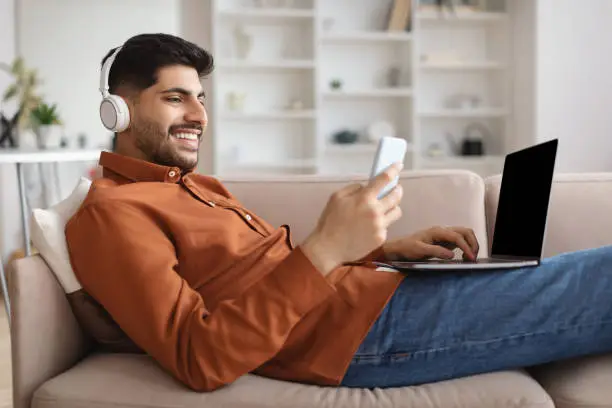 Photo of Smiling man in wireless headhones using cellphone and pc