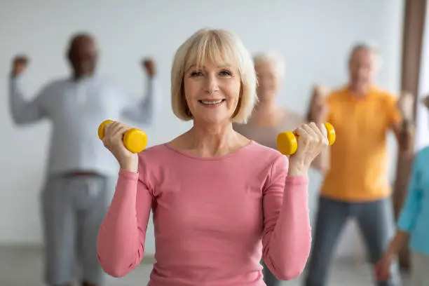 Selective focus on cheerful senior lady posing with fitness tools, multiracial group of healthy elderly people in sportswear doing strength building workout with dumbbells at gym