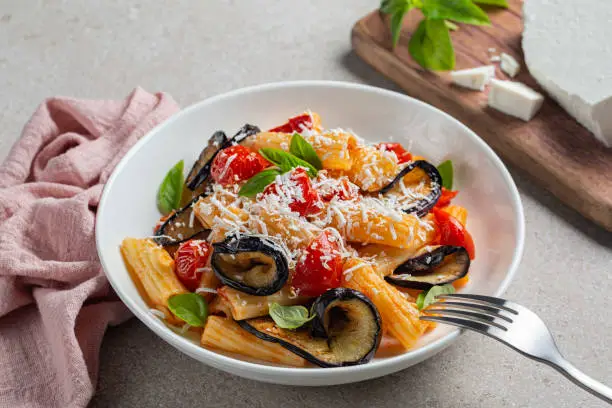 Pasta penne with eggplant, tomato sauce, basil, served with grated ricotta salata cheese. Pasta alla Norma, pasta salad. Pink and beige table surface. Close-up.