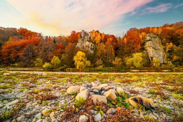 High rocky cliffs among lush yellowed trees growing near mountain river with clear water in autumn under blue sky and pink clouds