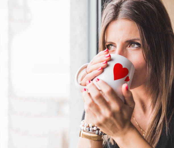 istock girl looking through a window while holding a white mug with red hearts. She is facing her and the light shines on her. The light is golden and soft. Rest time. She is in love. 1368999807
