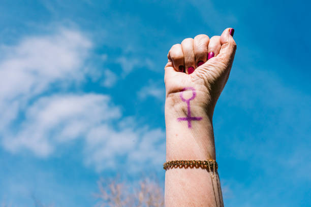 Fist of an older woman with purple painted nails, with the sky in the background, with the female symbol painted. Concept of women's day, empowerment, equality, inequality, activism and protest. stock photo