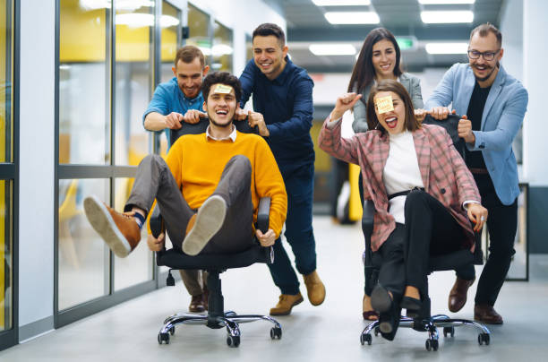 jovens empresários alegres se divertindo enquanto correm em cadeiras de escritório e sorrindo. - office fun competition chair - fotografias e filmes do acervo