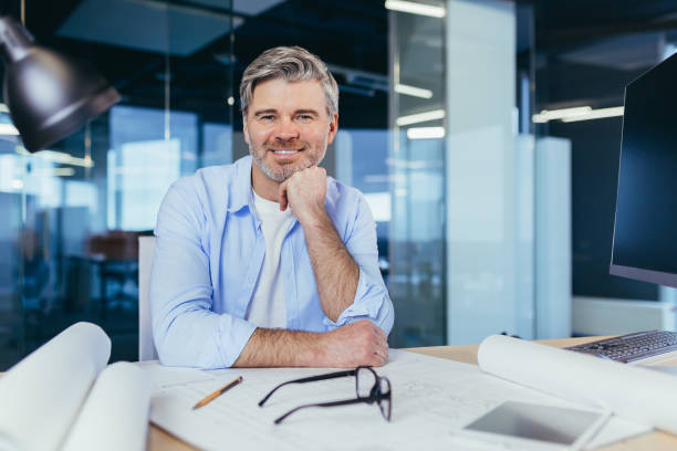 istock Experienced gray-haired designer architect, smiling and looking at camera, man working on architectural projector of new office center 1369002862