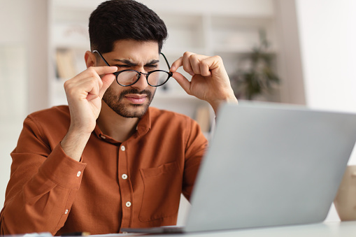 Young male freelancer with bad eyesight using laptop, trying to work from home. Middle Eastern guy holding and taking off his glasses and squinting, looking at laptop screen, having vision troubles