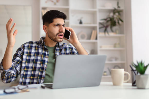 istock Confused Arab man talking on mobile phone using laptop 1369004772
