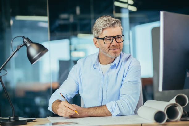 istock Experienced designer architect works at the table draws a draft plan of the house, a man looks at the monitor, a man at work 1369005515