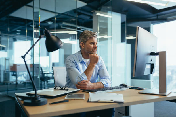 istock A gray-haired man thinks about the tasks, a businessman with a beard works in a modern office at the computer, 1369005552