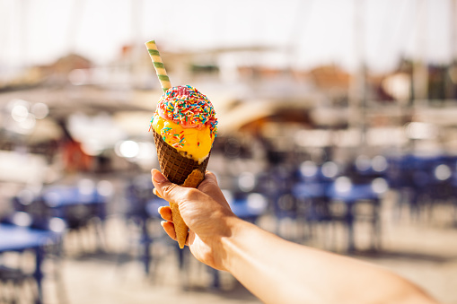 Young woman holding cone with beautiful ice cream. In the background are restaurants and marina with yachts and boats. She is tourist on vacation on Mediterranean island in Greece.