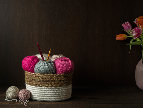 colorful woolen balls with crochet hooks in a wooden basket in front of a dark brown wooden background