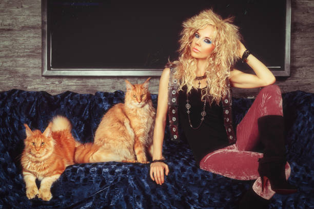 vintage-porträt einer schönen jungen blonden frau mit einer meeräschenfrisur im glam-rock-stil der 80er jahre, die auf einer mit einem blauen überwurf bedeckten couch posiert und neben zwei reinrassigen maine coon-katzen aus ingwer sitzt - heavy metal stock-fotos und bilder