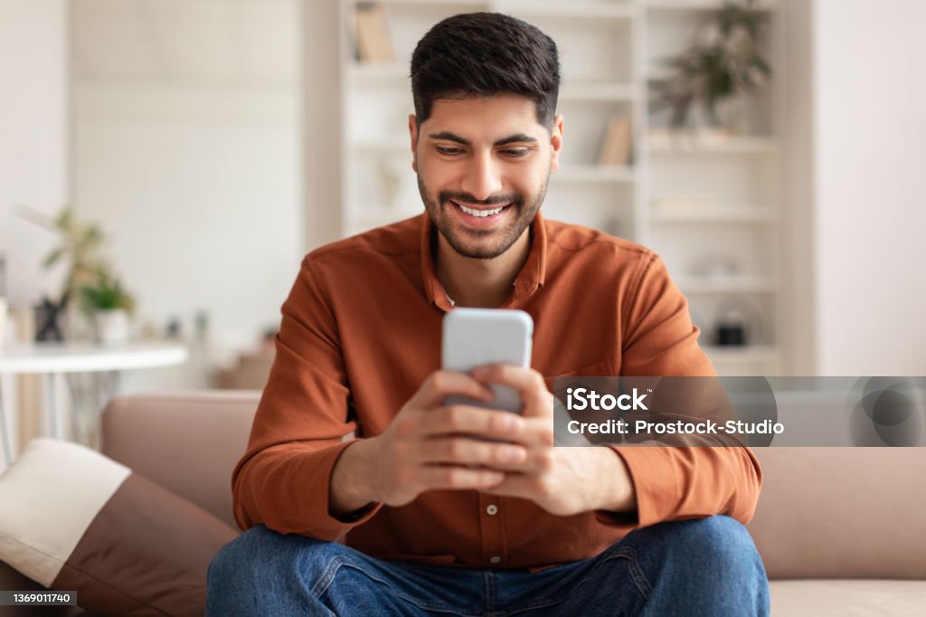 Portrait of smiling Arab man using smartphone at home Cool Gadget And Application. Portrait of young smiling Arab man holding mobile phone, typing sms message, sitting on the couch in living room. Guy browsing internet, surfing web, using app, free space Men Stock Photo