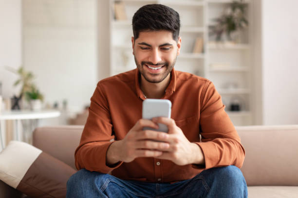 Portrait of smiling Arab man using smartphone at home Cool Gadget And Application. Portrait of young smiling Arab man holding mobile phone, typing sms message, sitting on the couch in living room. Guy browsing internet, surfing web, using app, free space arab culture stock pictures, royalty-free photos & images