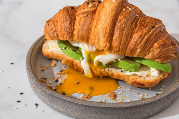 сэндвич с круассаном с яйцом пашот, авокадо и мягким сыром в тарелке на белом фоне. французский завтрак - croissant morning white breakfast стоковые фото и изображения