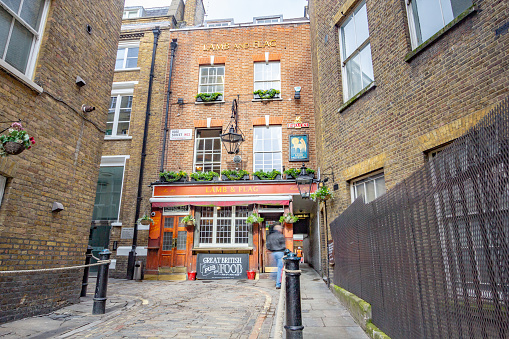 A person walking past commercial signs visible on Lamb & Flag which dates from the early 18th century, having become a pub in at least 1772. In the upper area, bare-knuckle prize fights took place so the pub was nicknamed 'the bucket of blood'. The pub was often visited by writer Charles Dickens frequented the pub in the 1800s and was resurfaced with brick in 1958.