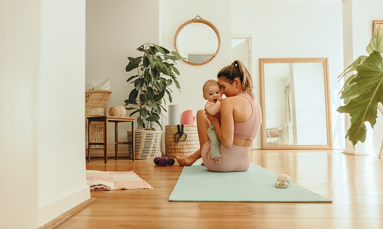 Affectionate mom working out with her baby at home. Healthy mom holding her baby while sitting on an exercise mat. New mom bonding with her baby during her post-natal fitness routine.