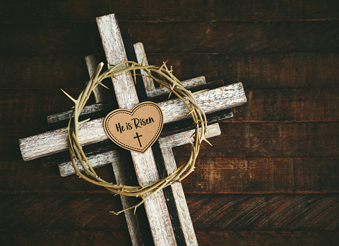 Wooden cross with a crown of thorns on a rustic wood background with a message that reads HE IS RISEN. Easter background