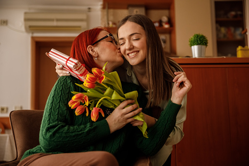 Front view of affectionate mature woman kissing and embracing her caring young daughter who is sharing a beautiful bouquet of tulips and  a gift for Mother's day.