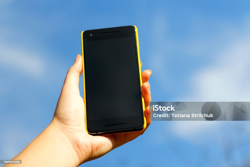 Woman hand holding black smartphone in yellow plastic case  on blue sky nature background, sunny day. Black screen. Mockup, mock-up, mock up. Female hand. Smart phone Woman hand holding black smartphone in yellow plastic case  on blue sky nature background, sunny day. Black screen. Mockup, mock-up, mock up. Female hand. Smart phone. Backgrounds Stock Photo