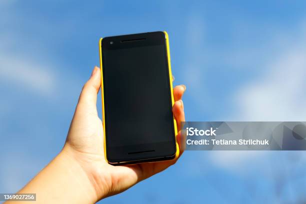 Woman Hand Holding Black Smartphone In Yellow Plastic Case On Blue Sky Nature Background Sunny Day Black Screen Mockup Mockup Mock Up Female Hand Smart Phone Stock Photo - Download Image Now