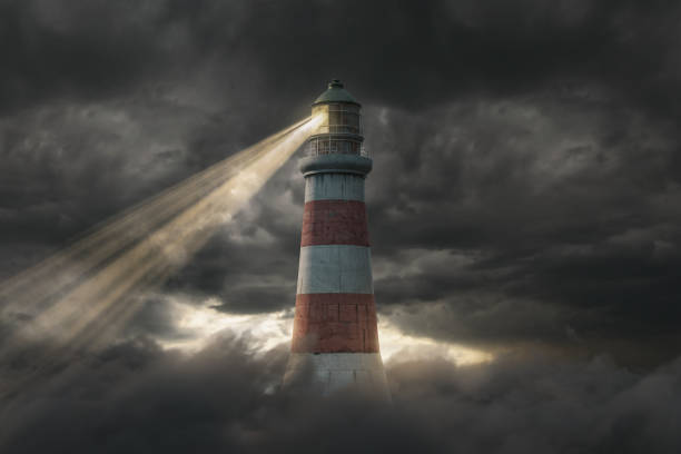 Lighthouse shining its lantern from a shoreline against a dramatic sky at dusk