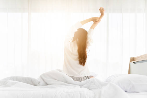 buongiorno nuovo giorno. la donna asiatica si sveglia e il corpo seduto si allunga sul letto accanto alla finestra in camera da letto - mattino foto e immagini stock