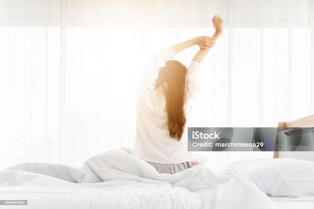 Good morning new day. Asian woman wake up and sitting body stretch on bed beside window in bedroom Waking up Stock Photo