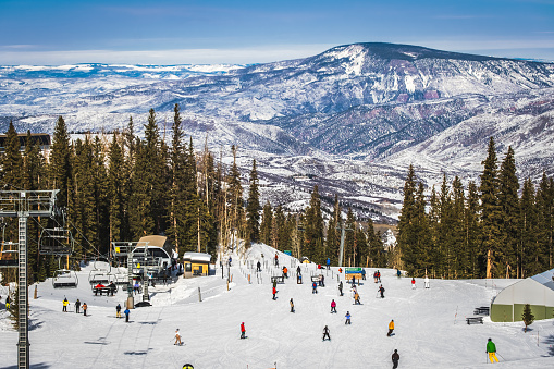 View of Colorado ski slope on clear winter day; people skiing and snowboarding to base of chairlift; forest and mountains in background