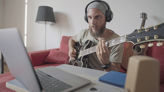 Online guitar teacher in video chat look at laptop talk and teach guitar during videoconference at home office