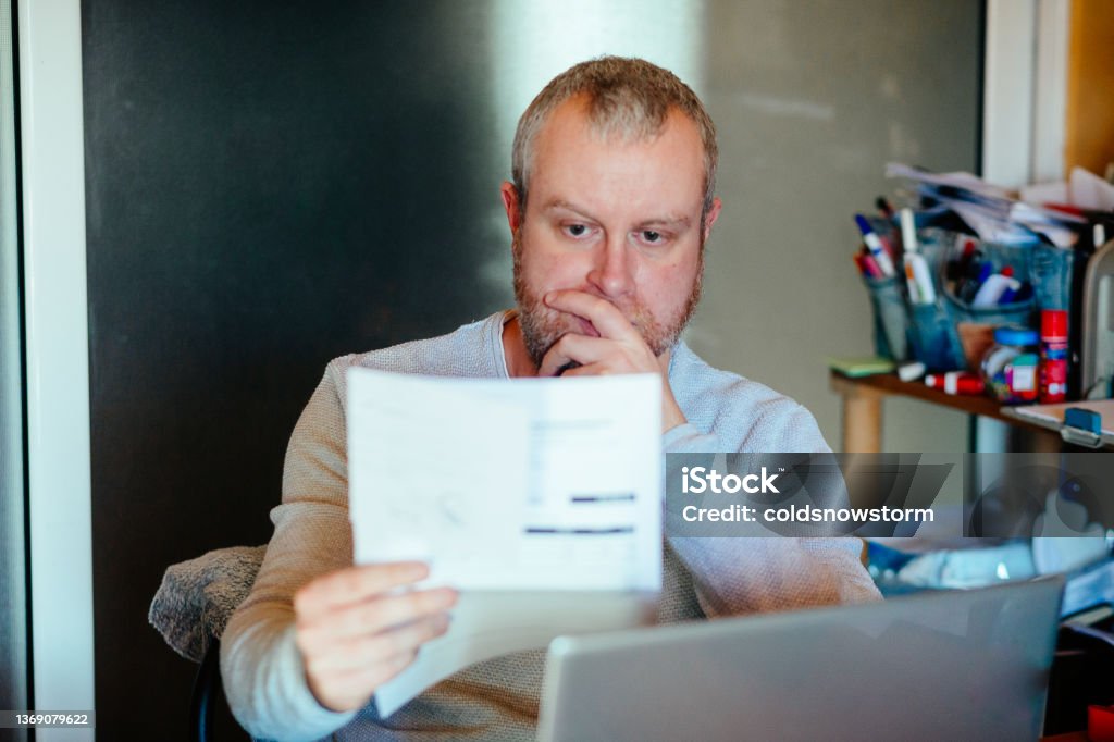 Worried man checking bills at home Portrait of a mid adult man checking his energy bills at home. He has a worried expression and touches his face with his hand while looking at the bills. Focus on the man while the interior architecture of the house is defocused. Financial Bill Stock Photo