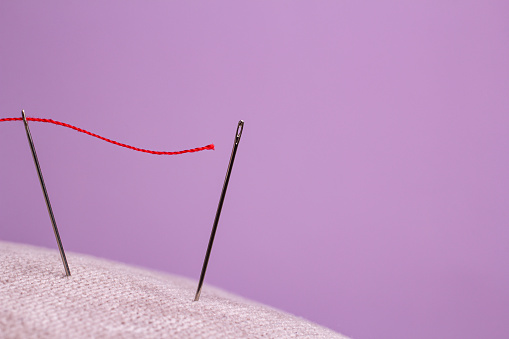 red thread for sewing is threaded into the eye of the needle and directed to the eye of the second needle on a pink background