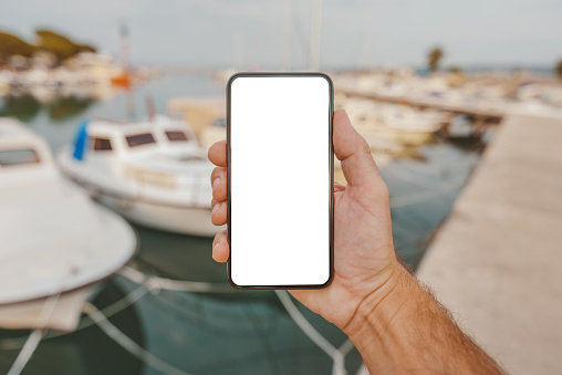 Fisherman holding smartphone with blank mock up screen in front of his boat in marina, selective focus