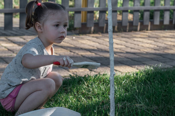 petite fille de 2-4 ans avec brosse blanchissant à la chaux la base du jeune tronc d’arbre dans le jardin - child caucasian little girls 3 4 years photos et images de collection