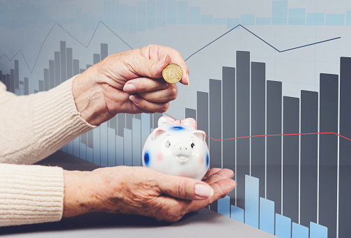 Wrinkled hands of an elderly woman puts a euro coin in a piggy bank. Piggy bank is defocused. Business chart on the background. The concept of savings and economy.