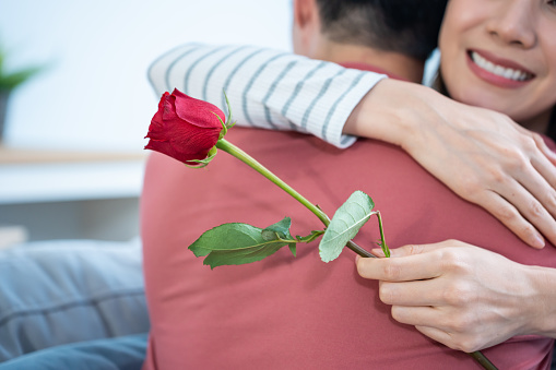 Asian young man surprise and give rose flower to beautiful girlfriend. Attractive romantic new marriage couple male and woman spend time to celebrate anniversary and valentine's day together in house.
