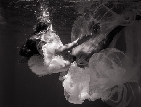 Beautiful young woman in bridal dress swims and dives under sunlit deep water with her reflection. Black and white image. Closeup.