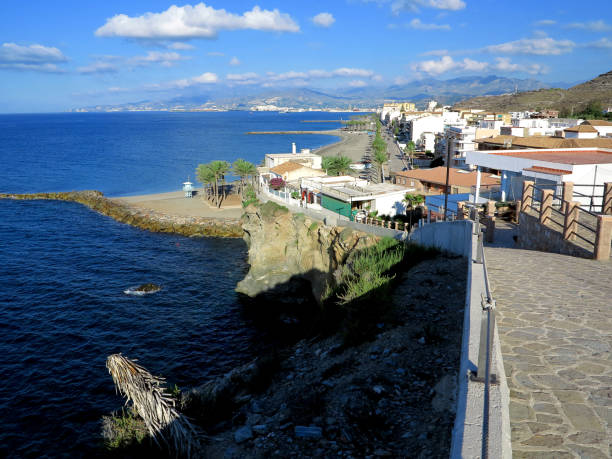 Stone paved cliff walking path above the Mediterranean sea with the town of Torrenueva Costa and mountains in the background with big blue sky and puffy white clouds.