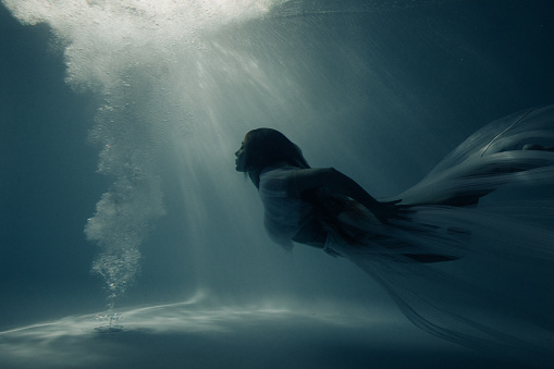 Beautiful woman in bridal dress swims and dives under sunlit deep water.
