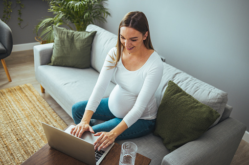 A pregnant woman in casual clothes is sitting and using a laptop on the sofa. Expecting a baby. Home atmosphere. Searching for pregnancy information.
