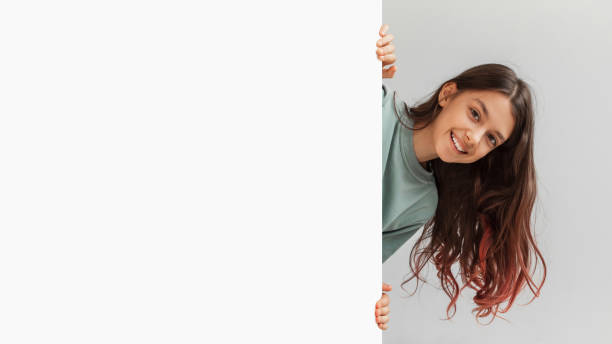 Happy Girl Hiding Behind Empty White Board Over Gray Background Happy Preteen Girl Posing Hiding Behind Empty White Board And Smiling To Camera In Studio Over Gray Background. Child Holding Paper Poster Advertising Your Text. Panorama, Mockup peeking stock pictures, royalty-free photos & images