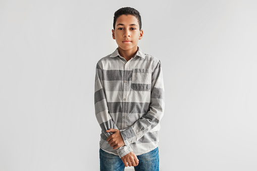 Serious Black Preteen Boy Looking At Camera Standing Over Gray Studio Background, Wearing Casual Clothes, Shirt And Jeans. Kids Fashion And Style Concept. Front View Shot