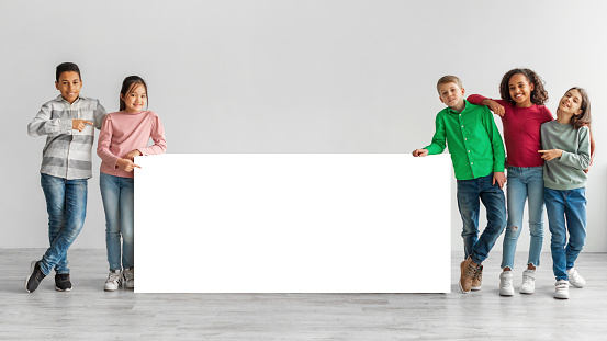 Group Of Happy Diverse Kids Standing Near White Blank Board Advertising Your Text Pointing Fingers Over Gray Studio Background. Boys And Girls Posing Together Near Paper Poster. Panorama, Mockup