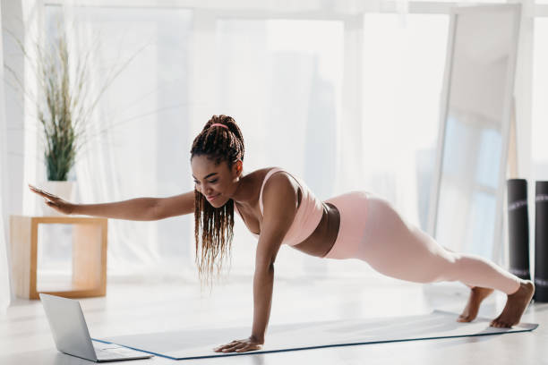 istock Fit young black woman standing in plank with outstretched arm, following online video tutorial on laptop at home 1369112736