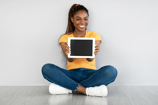 Beautiful black woman showing tablet computer with empty screen, sitting on floor against grey wall, mockup for website or application. African American lady demonstrating touch pad display template