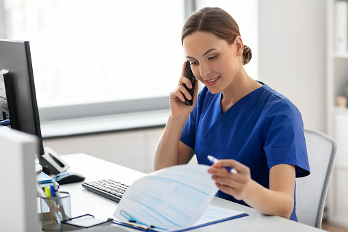 medicine, technology and healthcare concept - happy smiling female doctor or nurse with computer and clipboard calling on phone at hospital